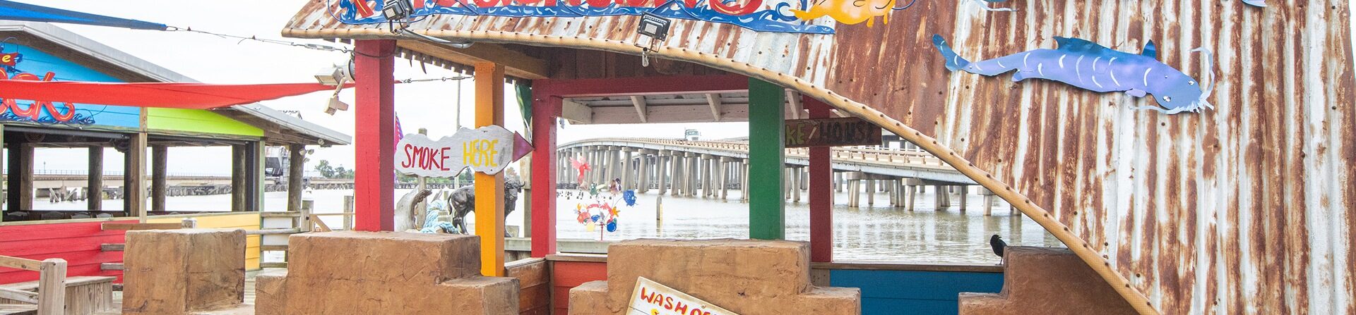Colorful, rustic outdoor dining area with corrugated metal roof, wooden beams, and playful sea-themed decorations, overlooking water and a bridge in the background.