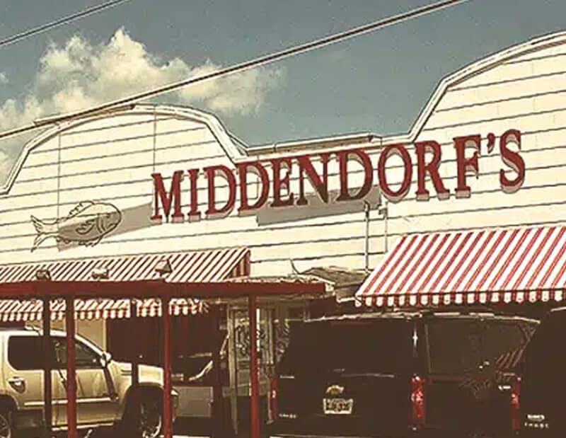 Exterior view of Middendorf’s restaurant with red striped awnings, a large sign, and several vehicles parked in front.