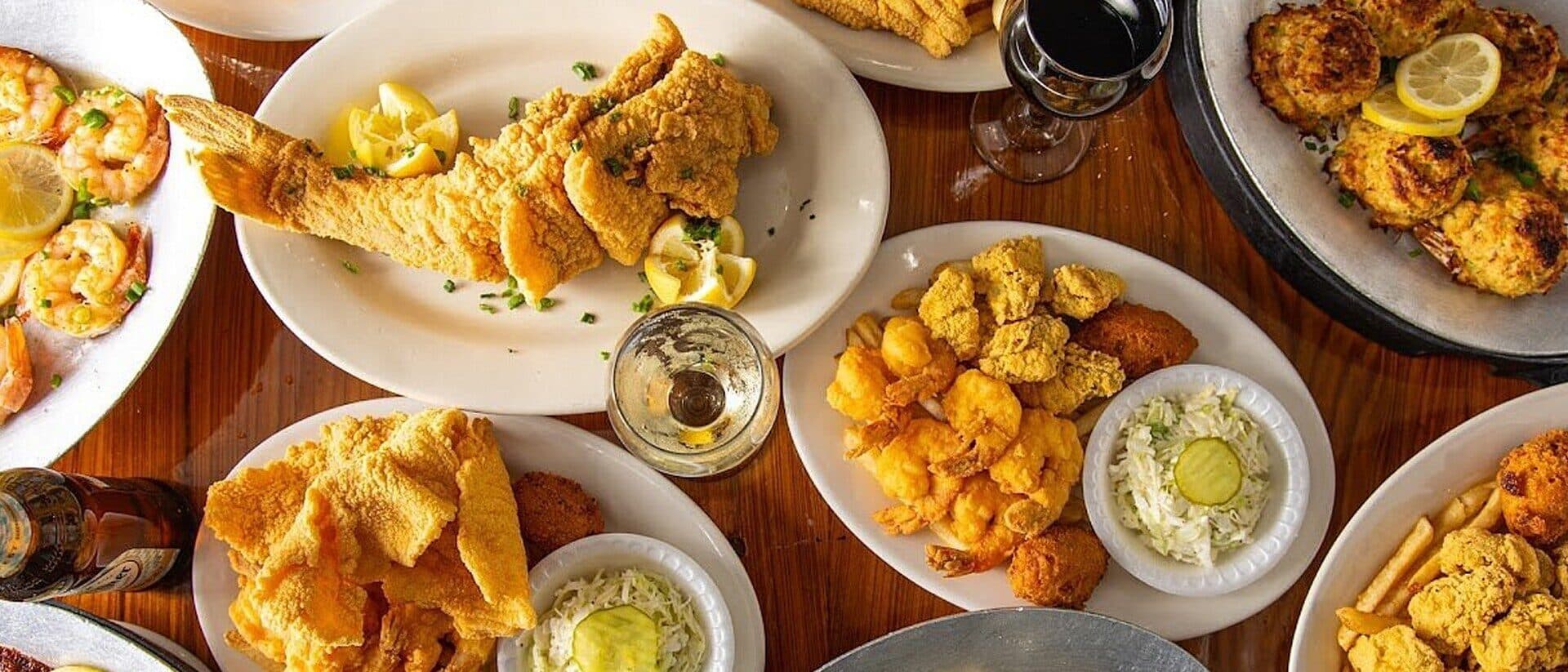 A table set with plates of fried fish, fried plantains, sliced onions, and dipping sauces, along with drinks.