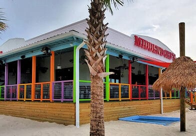 A colorful, tropical-themed beachfront restaurant with a raised, wraparound porch, palm trees, and a thatched umbrella on sandy ground.