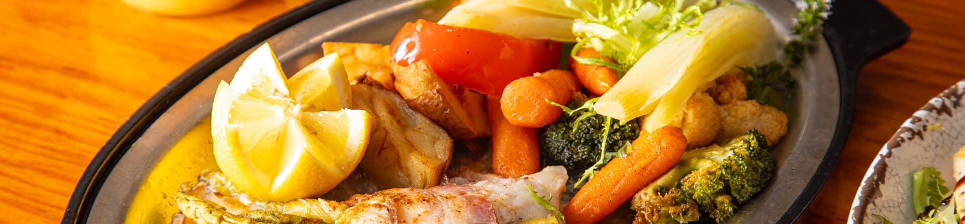 A sizzling platter of grilled fish, seasoned vegetables, and lemon wedges on a black plate, served on a wooden table.