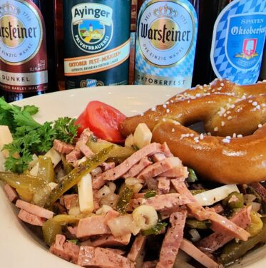 A plate with a sliced sausage salad, pretzel, greens, and tomato sits in front of five assorted bottled beers on a wooden table.