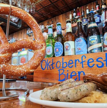 A large pretzel, plate of sausages, and sauerkraut are displayed on a bar counter with various beer bottles and an "Oktoberfest Bier" sign in the background.