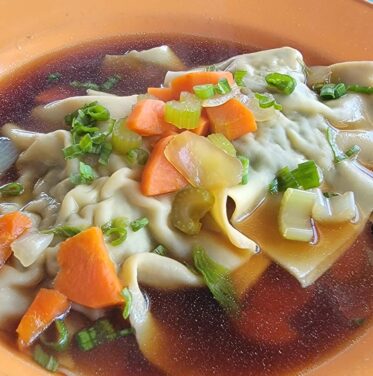A plate of dumplings in clear broth, garnished with chopped carrots and green onions, with two spoons on the side.
