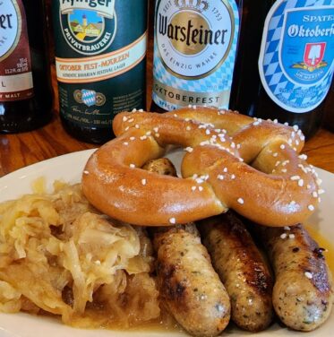 Plate with three grilled sausages, sauerkraut, and a soft pretzel in front of assorted bottles of German beer on a wooden table.