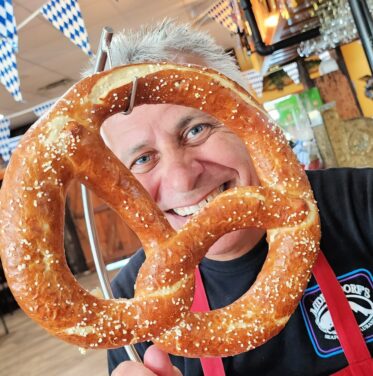 A man holds a large pretzel in front of his face inside a restaurant decorated with blue and white checkered flags.