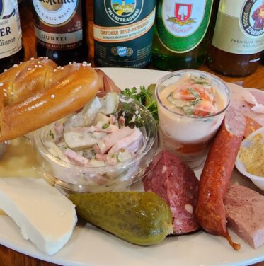 A plate with assorted sausages, sliced cheese, a pickle, mustard, a pretzel, cold salad, and a small glass of creamy salad, with bottled beers in the background.