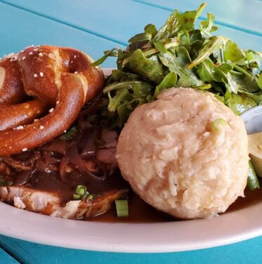 A plate of sliced roast meat with gravy, a large bread dumpling, a soft pretzel, mustard, and arugula salad on a blue table—perfect for savoring during Oct to Nov-Oktoberfest celebrations.