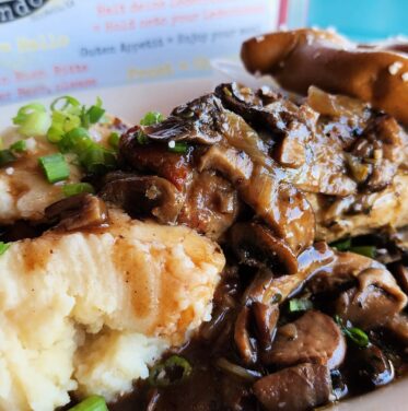 Plate of mashed potatoes and meat topped with mushroom gravy, garnished with green onions, served with a pretzel twist on the side—a hearty dish perfect for Oktoberfest celebrations from Oct to Nov.