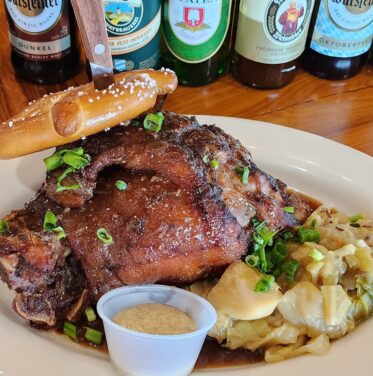 A plate of roasted pork knuckle garnished with green onions, served with sauerkraut, dumplings, a pretzel, and mustard—paired with assorted beer bottles in the background for the perfect Oct to Nov Oktoberfest feast.