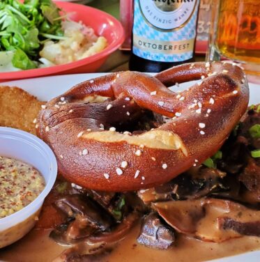 A plate with a soft pretzel, breaded appetizer, mustard dip, and meat in sauce—paired with a side salad and beer—sets the perfect Oktoberfest scene from Oct to Nov. A beer bottle and glass complete the festive background.