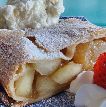 A slice of apple strudel dusted with powdered sugar, served with whipped cream, a strawberry, and a dollop of cream on a blue plate—a perfect treat for Oct to Nov Oktoberfest celebrations.