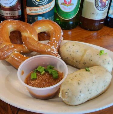 A plate with a soft pretzel, two breaded rolls, a cup of dipping sauce topped with green onions, and assorted beer bottles in the background.