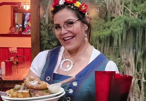 A woman wearing glasses and a flower headband smiles while holding a plate of food and red cups in a restaurant setting.