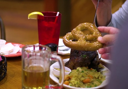 A person holds a soft pretzel above a plate of food, with a mug of beer and a red glass of iced drink on the table.