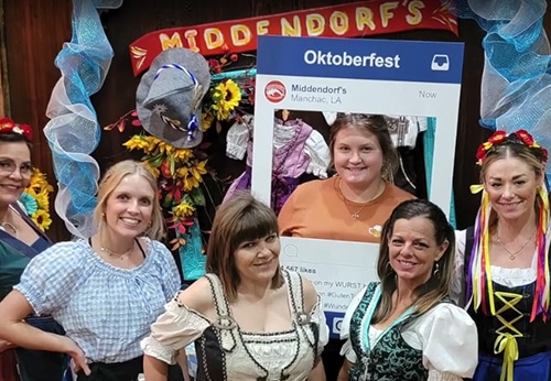 Five women in traditional German dresses pose in front of an Oktoberfest sign and a photo prop frame at an indoor event decorated with flowers and hats.