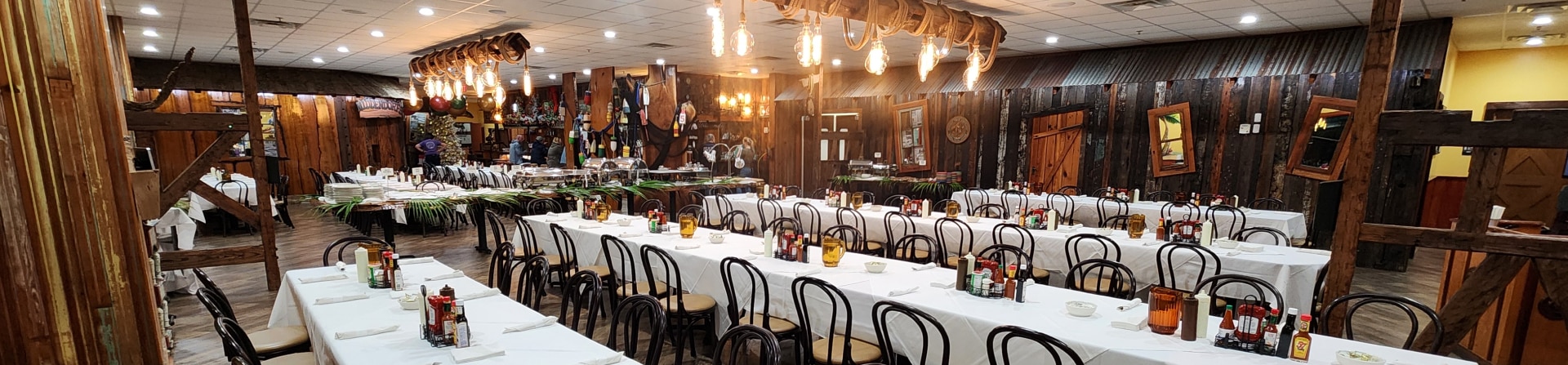 Large dining hall with long tables covered in white tablecloths, wooden chairs, rustic decor, and warm overhead lighting.