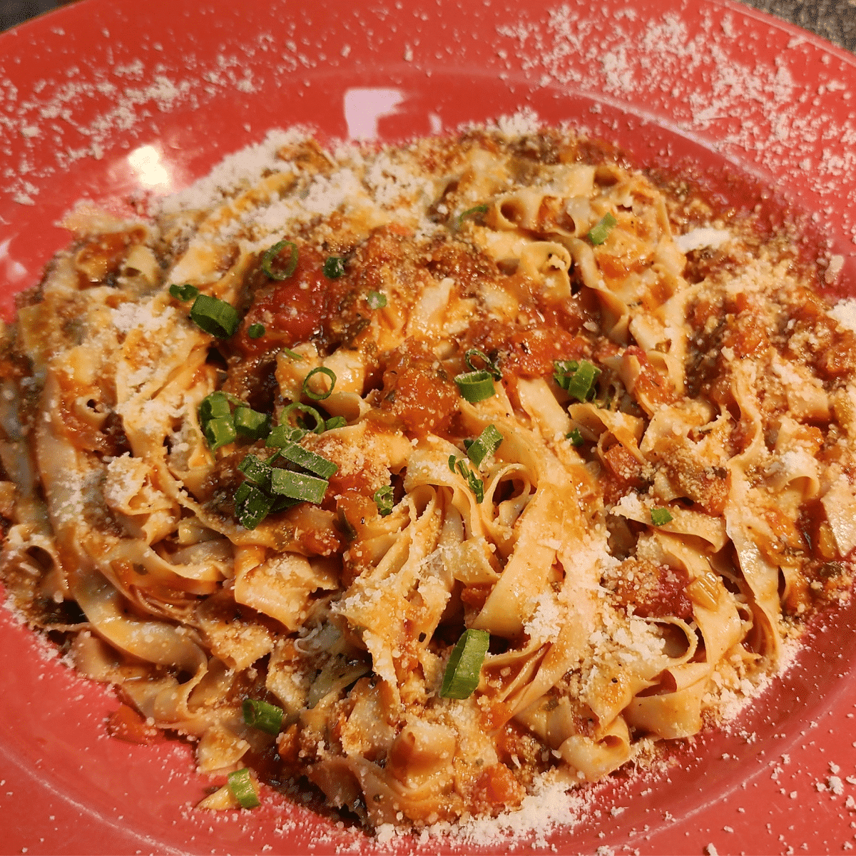 A plate of tagliatelle pasta topped with tomato sauce, grated cheese, and chopped herbs, served on a red dish.