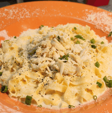 A plate of fettuccine pasta topped with shredded crab meat, grated cheese, chopped green onions, and cracked black pepper.